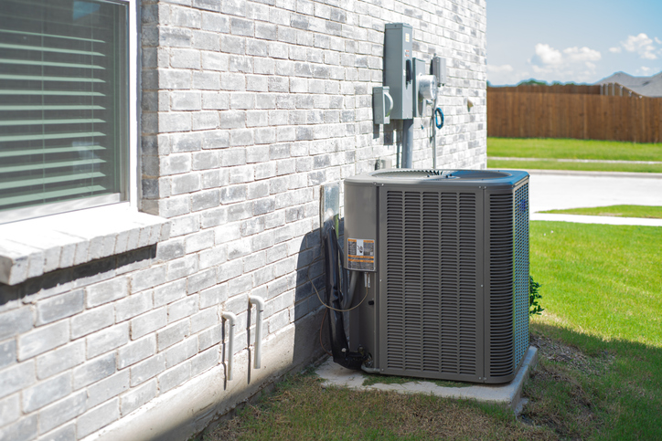 Outdoor residential HVAC unit installed beside a modern brick house. The unit sits on a concrete pad next to utility meters and piping, with a well-maintained lawn and clear blue sky in the background.