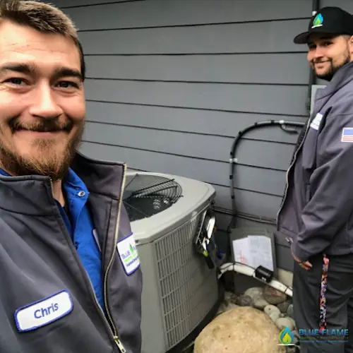 Two Blue Flame HVAC technicians standing next to an air conditioning unit outside a home, smiling for a selfie while working on the system.