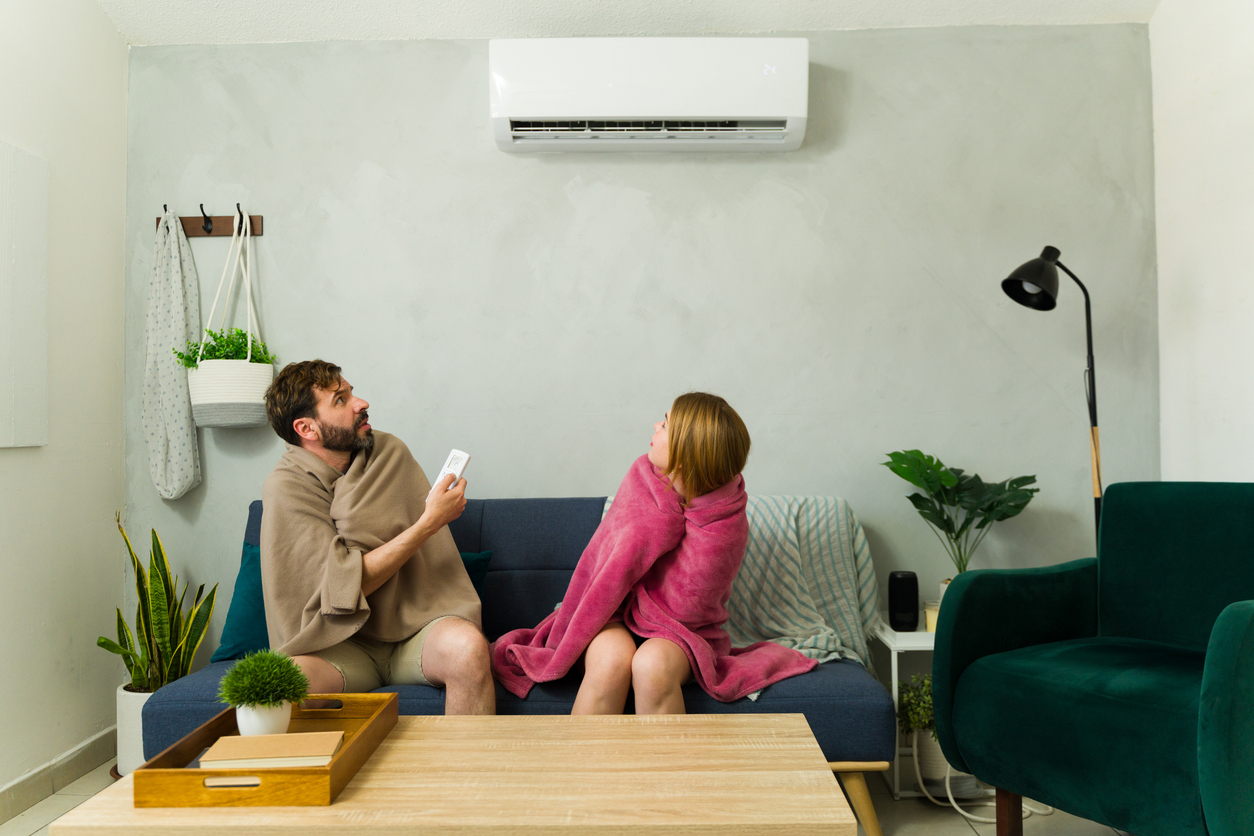 A man and a woman wrapped in blankets, freezing and looking up at the air conditioner.